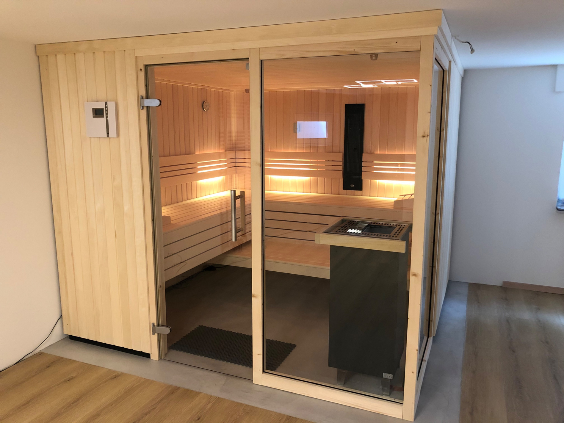 Sauna in Kellerraum, raumhoch, mit Eckverglasung und freistehendem Saunaofen. In der rückseitigen Saunawand ist ein Tiefnwärmestrahler als weiter Badeform eingebaut.